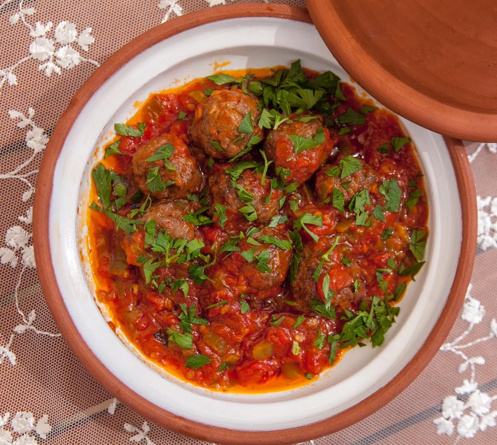Toprak güveçte domates soslu köfte, üstünde maydanoz; dantel örtü. Kare.