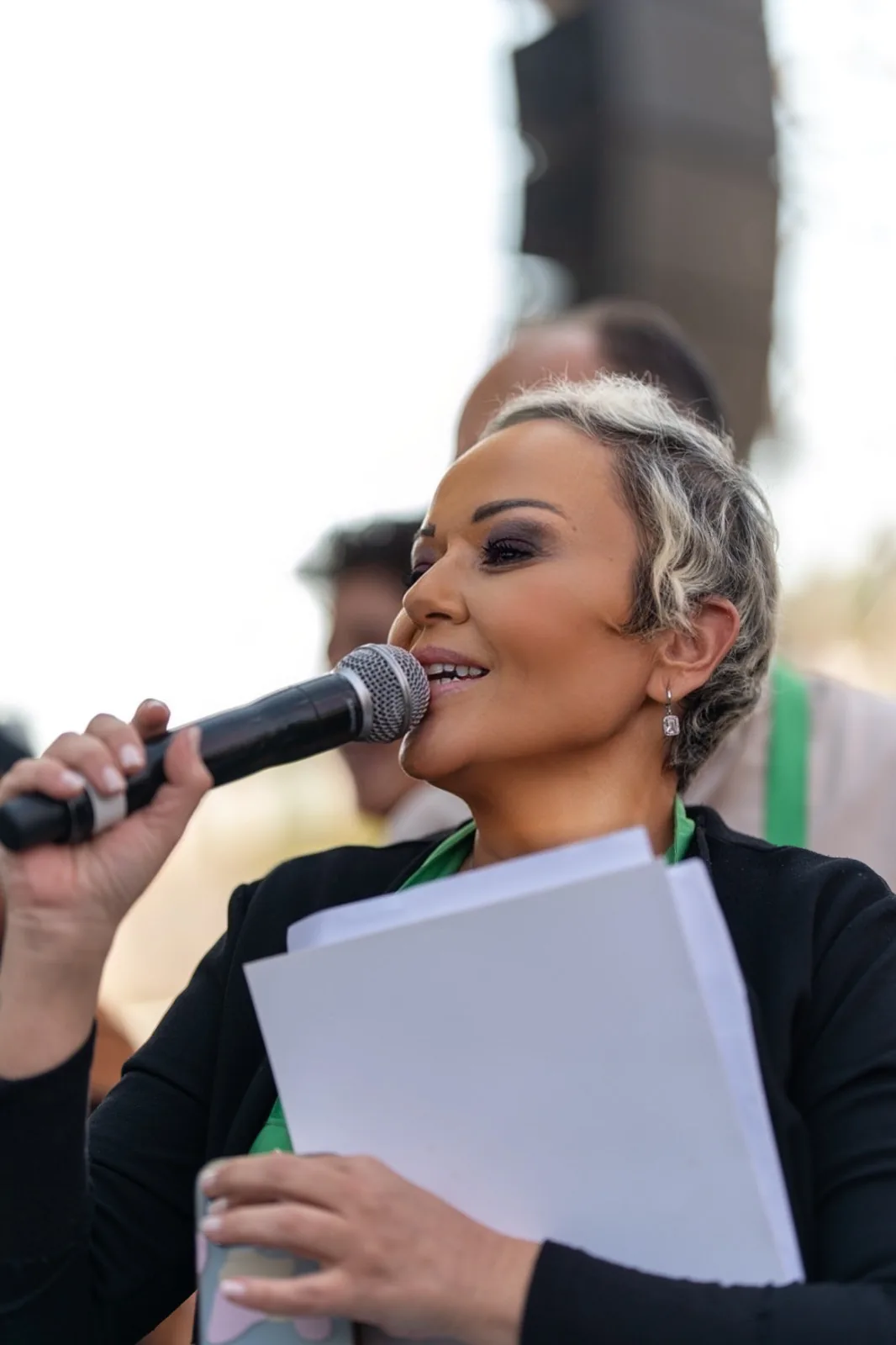 Yakın plan: Özlem kısa gümüş saçlarıyla mikrofonla konuşuyor, elinde akış kâğıtları; arka plan bulanık açık hava.