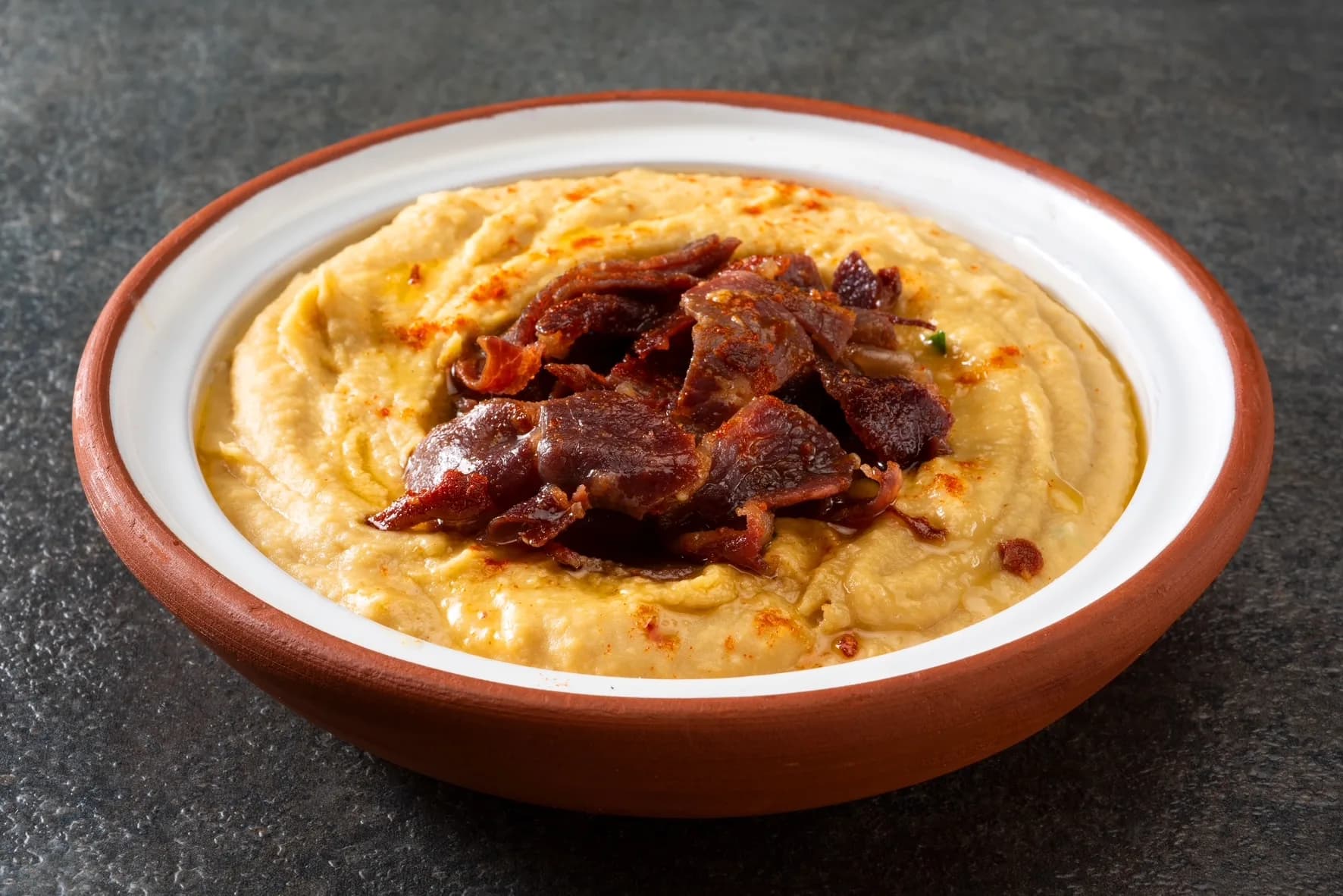 Toprak kenarlı kâsede humus, üstünde kızarmış et dilimleri ve pul biber. Yatay.