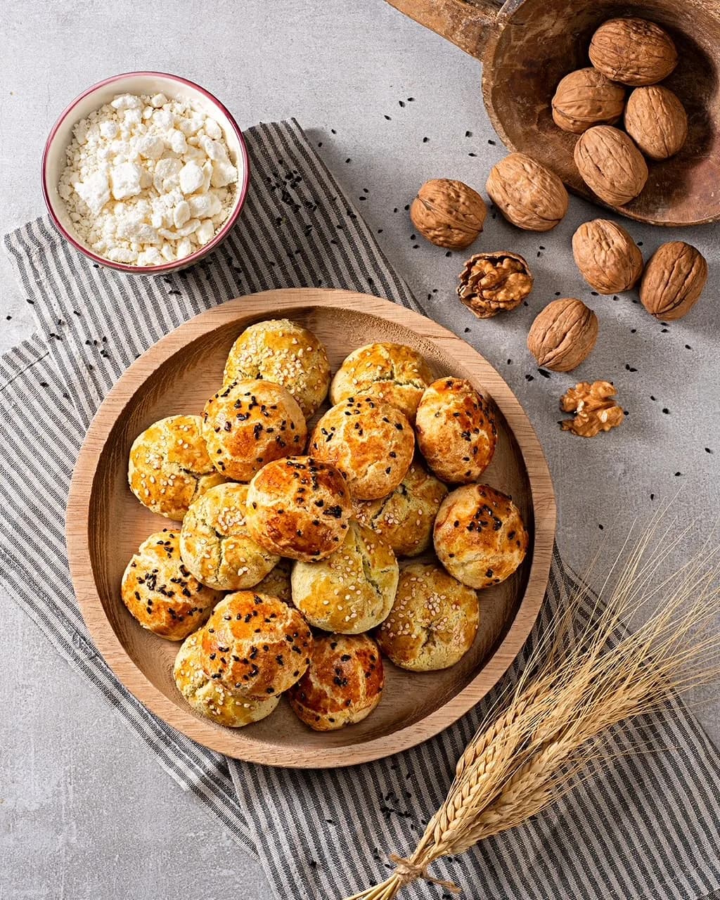 Ahşap tabakta çörek otlu peynirli poğaçalar; yanında ceviz, beyaz peynir ve başak. Kuşbakışı, dikey.
