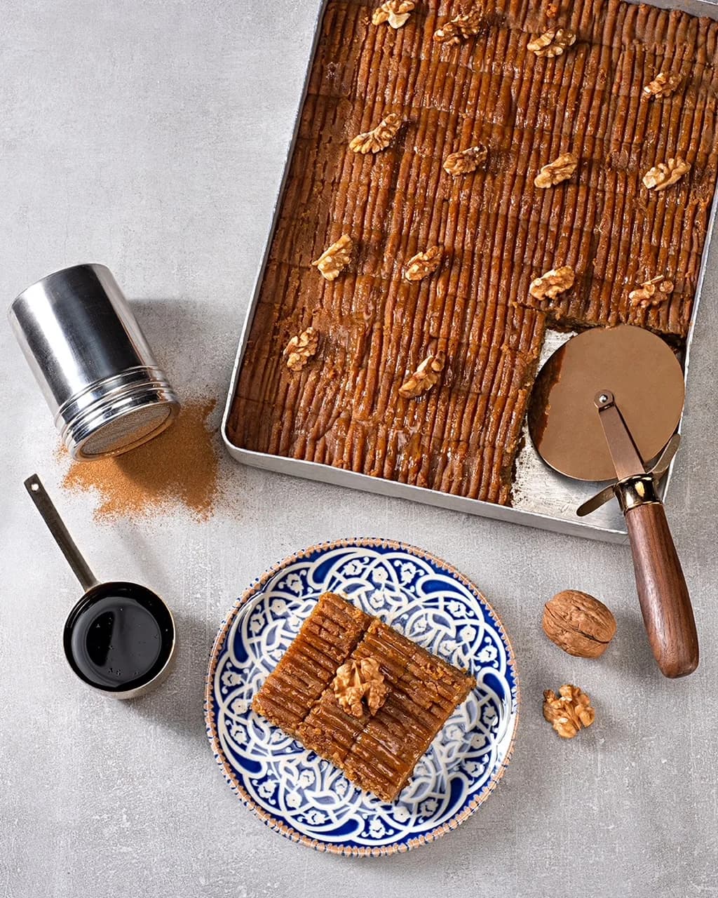 Tepside cevizli baklava, desenli tabakta bir porsiyon; yanında tarçın kutusu ve baklava kesme aleti. Kuşbakışı, dikey.