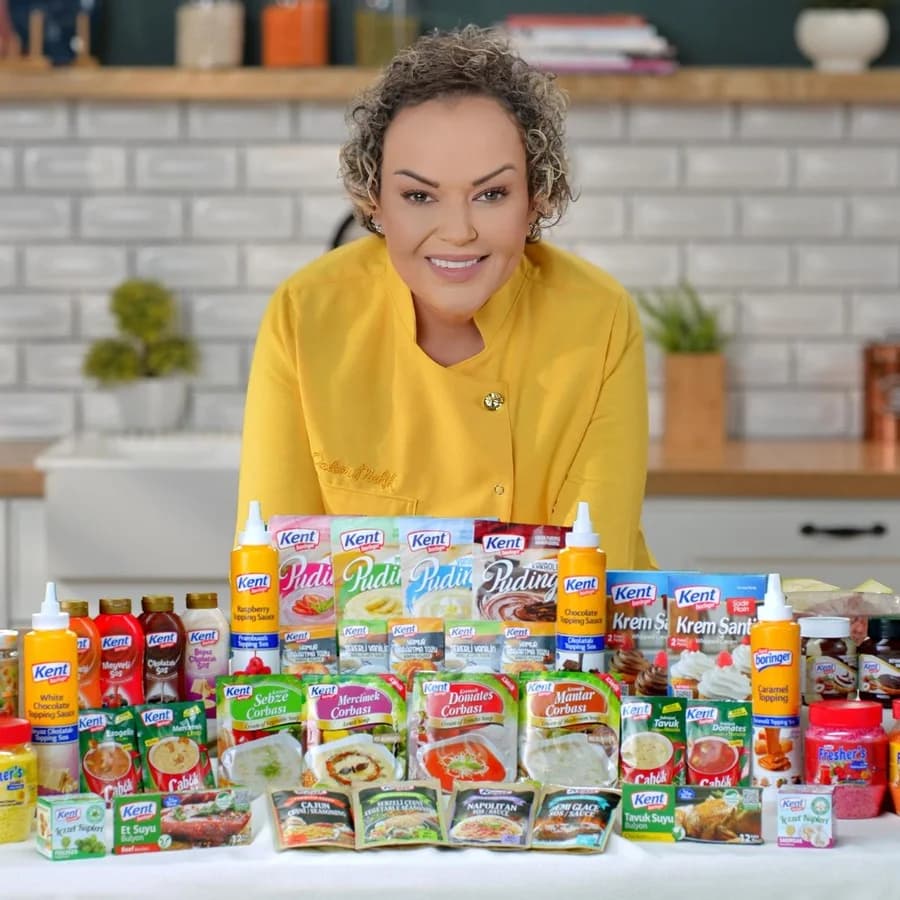 Kent ürün duvarı: Özlem sarı şef ceketiyle beyaz fayanslı bir mutfakta, önünde tezgâha dizilmiş Kent paketleri (puding, krem şanti, sos şişeleri, toz karışımlar); kameraya gülümsüyor. Kare.