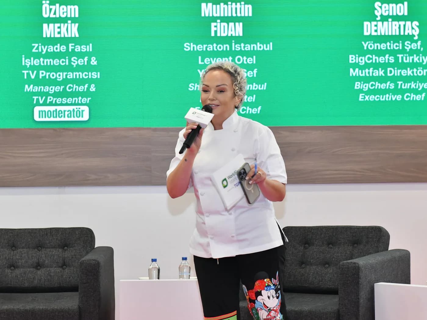 Panel sahnesi: Özlem beyaz şef ceketiyle elinde mikrofon ve kartlarla ayakta konuşuyor; arkadaki yeşil ekranda üç konuşmacının adı, kendi adının altında 'Ziyade Fasıl İşletmeci Şef & TV Programcısı · moderatör'. Yatay.