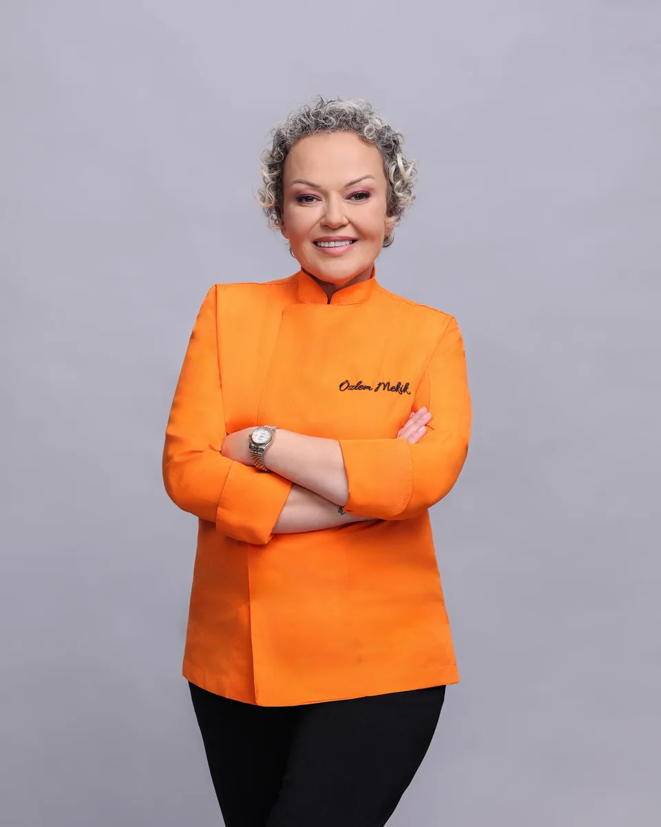 Özlem Mekik turuncu şef ceketi ve siyah pantolonla, kolları kavuşturulmuş, kameraya gülümsüyor