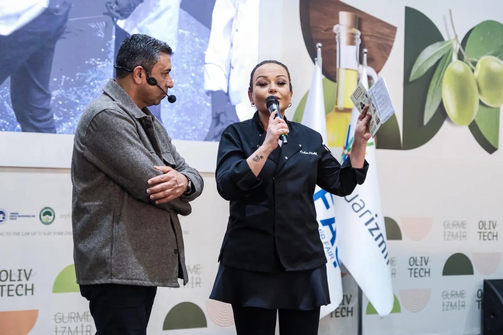 İzmir OlivTech fuarının sahnesi: Özlem siyah şef ceketiyle mikrofonla konuşuyor, elinde soru kartı; yanında kafa mikrofonlu, gri ceketli bir konuk dinliyor.