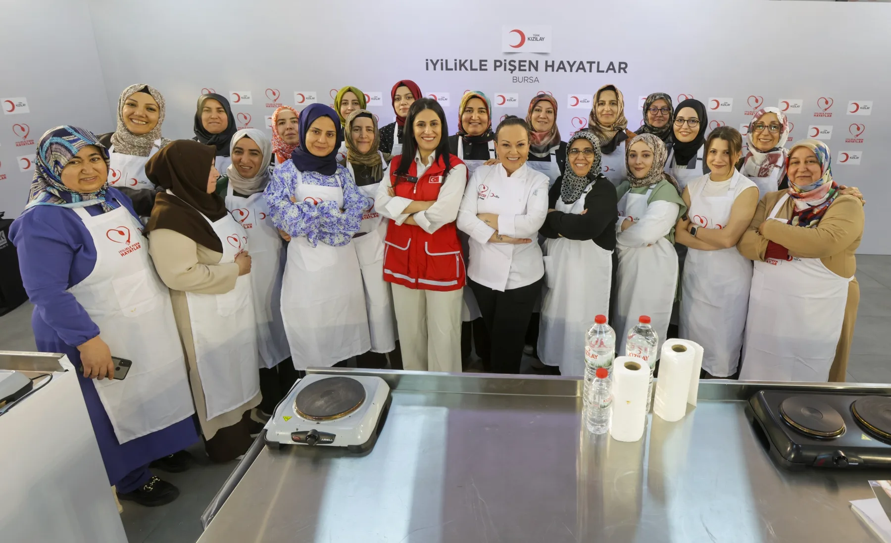 Aynı Kızılay eğitiminin toplu fotoğrafı: beyaz önlüklü kadın katılımcılar yan yana, ortada Özlem beyaz şef ceketiyle, yanında kırmızı Kızılay yelekli bir görevli; önde çelik tezgâhta elektrikli ocaklar.
