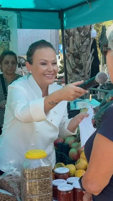 Çadırlı bir pazar tezgâhı: Özlem beyaz şef ceketiyle mikrofonu tezgâhtaki birine uzatıyor; önde kavanozlar ve meyve.