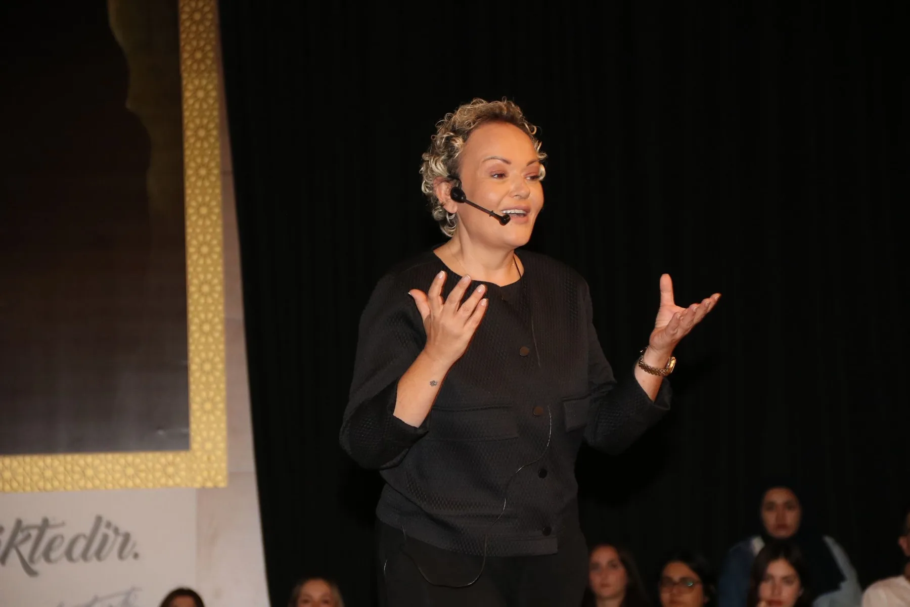 Siyah perdeli, altın çerçeveli bir salon: Özlem kafa mikrofonuyla iki elini açmış anlatıyor; önde dinleyen seyirci başları görünüyor.