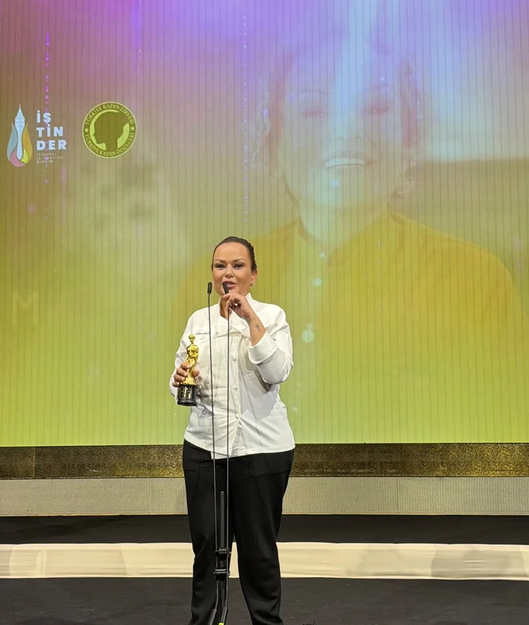 Aynı gece: Özlem beyaz şef ceketiyle sahnede mikrofon başında konuşuyor, elinde altın heykelcik; arkada büyük ekranda kendi görüntüsü. Dikey.