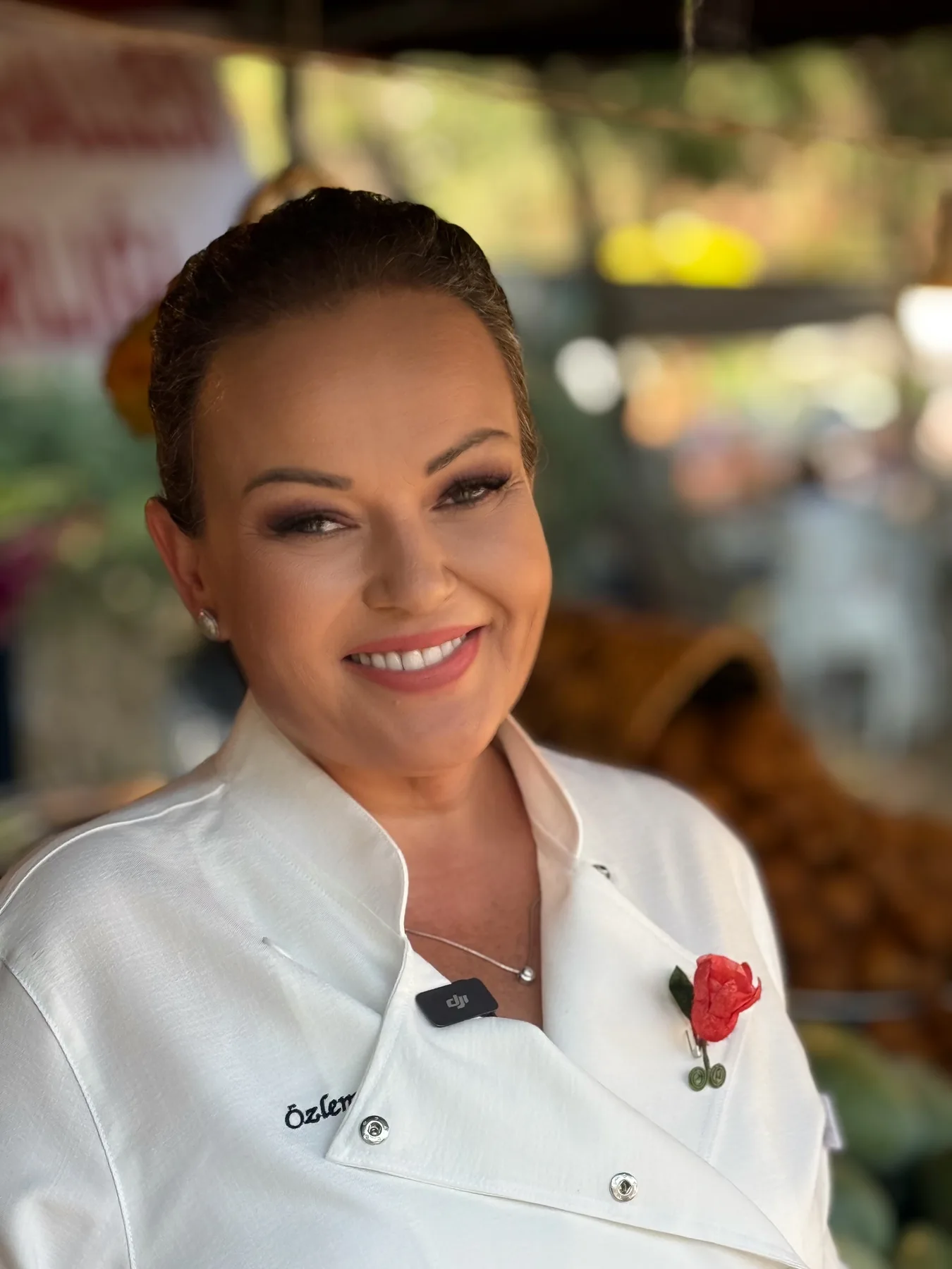 Festivalde yakın portre: Özlem beyaz şef ceketiyle (yakada kırmızı gül) gülümsüyor, arka plan bulanık yeşillik.
