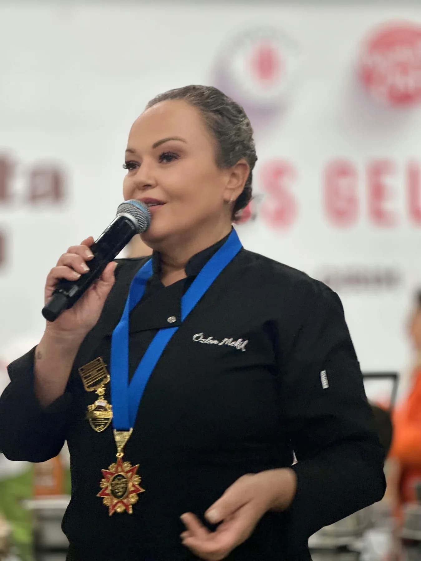 Özlem siyah şef ceketiyle, boynunda kurdeleli nişan, mikrofonla konuşuyor; arkada kırmızı beyaz festival pankartı.