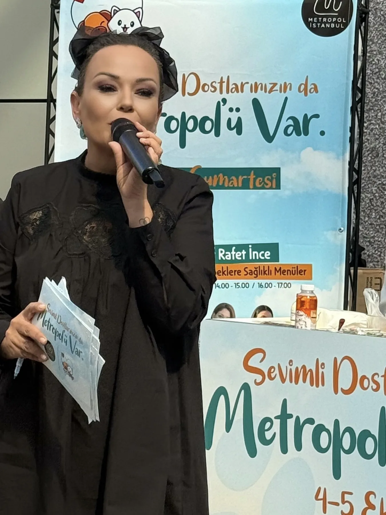 'Sevimli Dostlarımızın da Metropol'ü Var' afişinin önünde Özlem siyah kıyafetle mikrofonla konuşuyor, elinde akış kartları.