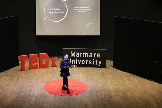 TEDx Marmara University sahnesi, uzak çekim: Özlem lacivert kıyafetle kırmızı halı dairenin üstünde konuşuyor; arkada slayt ve 'Marmara University' yazısı.