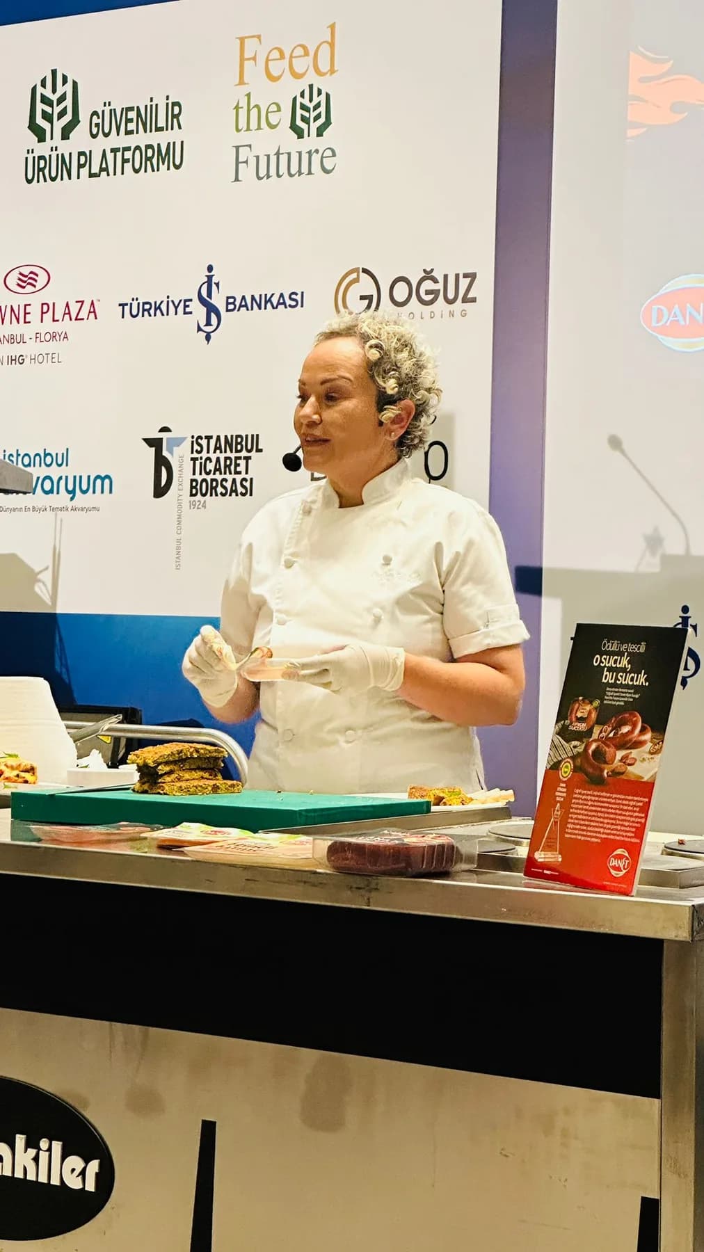 Aynı zirvenin gösteri tezgâhı, yakın: Özlem eldivenli elleriyle anlatıyor; önde yeşil kesme tahtasında dilimlenmiş börek. Dikey kare.